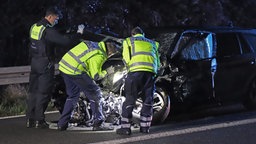 Polizisten stehen mit dem Rücken zur Kamera und begutachten einen Unfallwagen.