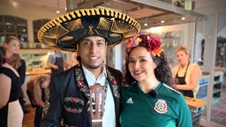Musiker Jean Carlos Giraldo Ante in einem traditionellen mexikanischen Musikeroutfit und Ledya Lopez von der Kochwerkstatt Düsseldorf im Fußballtrikot Mexikos.