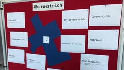 Eine rote Tafel mit weißen Zetteln mit den Ortsnamen, in der Mitte ist ein blaues Kreuz
