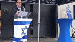 Zu sehen ist ein Redner an einem Rednerpult mit der Flagge Israels.