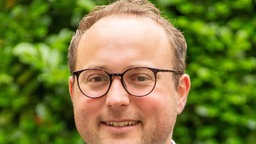 Ein Mann mit hoher Stirn, runder, markanter Brille und im Anzug vor grünen Bäumen
