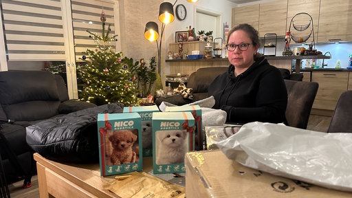 Nicole Schulz sitzt auf einem Sofa im Wohnzimmer, vor ihr steht ein Tisch mit Paketlieferungen.