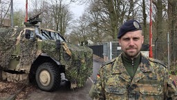Ein Mann in Soldatenuniform steht vor einer Einfahrt, hinter ihm ein Militärfahrzeug