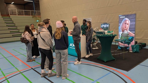 Eine Sporthalle mit einem Messe-Stand der AOK. Einige Mitarbeiter und eine Gruppe Schüler stehen um den Stand versammelt.
