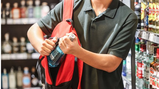 Zu sehen ist ein Jugendlicher, welcher eine Alkoholflasche in einen Rucksack steckt