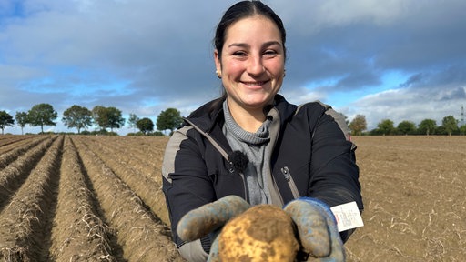 Eine junge Bäuerin steht auf einem Feld und lacht. Sie zeigt eine gerade aus dem Boden geholte Kartoffel.