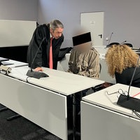 Der Angeklagte sitzt neben einer Dolmetscherin im Gerichtssaal. Auf seiner anderen Seite steht sein Verteidiger.