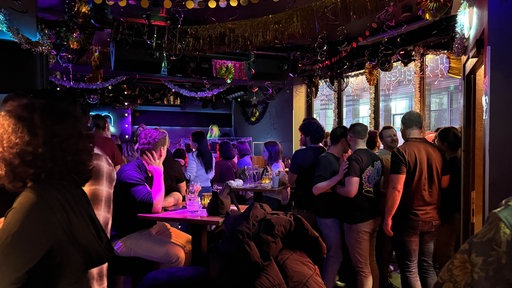 In einer Bar in köln stehen mehrere Personen und unterhalten sich, die Bar ist feierlich geschmückt, an der Decke hängen Glitzer-Girlanden.