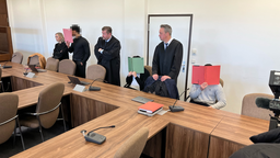 Drei Angeklagte halten sich Mappen vor das Gesicht, neben Ihnen stehen Anwälte in schwarzer Kleidung