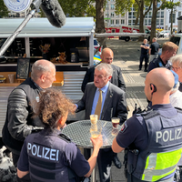 Herbert Reul steht mit zwei Polizisten und drei Männern um einen Tisch herum. Sie stehen vor einem Café-Wagen. Drumherum stehen weitere Polizisten und Polizeiwagen. Über den Tisch um den Herr Reul und die anderen stehen wird ein Mikrofon gehalten.