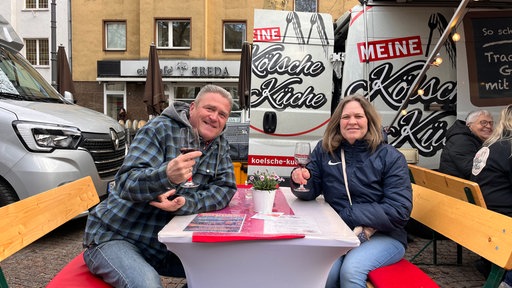 Zwei Gäste sitzen mit Rotweingläsern in der Hand auf dem Abendmarkt
