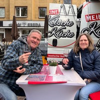 Zwei Gäste sitzen mit Rotweingläsern in der Hand auf dem Abendmarkt