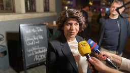 Berivan Aymaz (Grüne) steht draußen vor einem Gastronomie-Schild. Ihr werden Mikrophone von Radio Köln und WDR entgegengehalten