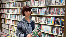 Eine Frau mit Pony-Frisur steht mit einem Buch in der Hand vor einem Bücherregal.