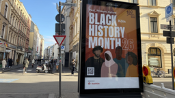 Im Vordergrund ist eine digitale Werbetafel, mit der Anzeige des Black History Month 26 zu sehen. Diese steht in der im Hintergrund zu sehenden Innenstadt 