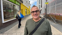Ein Mann steht in der Fußgängerzone und lächelt in die Kamera. er trägt einen olivgrünen PUlli und eine schwarze Brille.