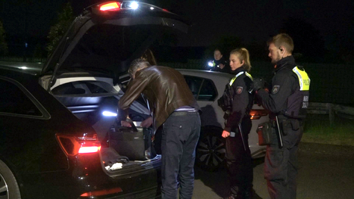 Ein Mann sucht etwas in seinem Kofferraum, daneben stehen zwei Polizisten