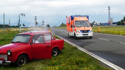 Ein Krankenwagen nähert sich einen verunfallten Trabi, aus welchem Rauch aufsteigt