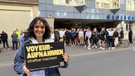 Die Kölnerin Yanni Gentsch, eine Frau mit dunklen Haaren und blauer Jeansjacke, steht vor einer Häuserfront mit einem Schild mit der Aufschrift "Voyeur-Aufnahmen strafbar machen"