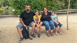 Eine Familie mit Mutter, vater und zwei Kindern sitzt im Zoo auf einer Bank im Sand
