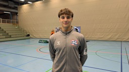 Handballer Luca Schulz steht in einer Sporthalle. Hinten ist ein Stand einer Jobmesse aufgebaut. Dahinter ist ein hallenfüllender weißer Vorhang.