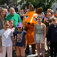 Einige Kinder und zwei Erwachsene stehen um eine orangene Mausfigur herum.