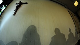 Schatten von Menschen an einer Wand, ebenso ein hängendes Kreuz