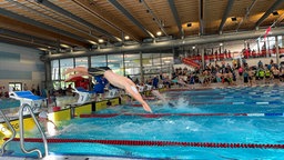 Ein Schwimmer mit weißer Badekappe springt mit einem Kopfsprung von einem Startblock ins Wasser