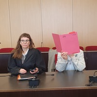 Der Angeklagte mit seiner Anwältin im Gericht. Er hält sich einen roten Ordner vor das Gesicht.