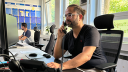 An einem Schreibtisch in einem Büro sitzt ein Mann mit Brille. In seiner rechten Hand hält er einen Telefonhörer.
