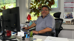 An einem Schreibtisch in einem Büro sitzt ein mittelalter Mann mit Brille. Seine rechte Hand liegt auf einer Computermaus. Seine linke Hand ist auf der Tastatur.