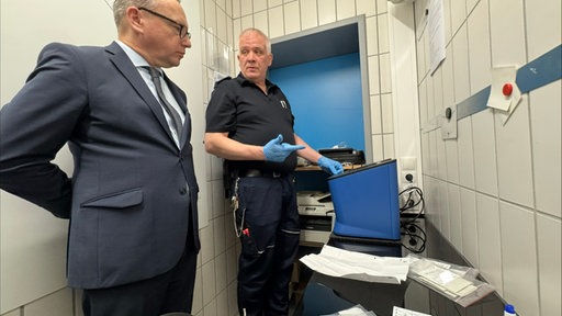 Ein Mann steht im Anzug neben einem Mann in blauer Gefängnis-Wärter-Kleidung. Auf dem Tisch vor ihnen steht ein blauer Kasten, so groß wie zwei Schuhkartons, und dort liegt ein Blatt Papier.