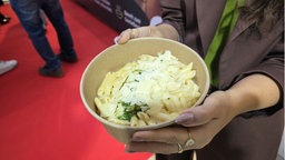 Auf dem Foto ist eine Frau, die eine Pappschale hält. Darin sind Nudeln mit Parmesan und Pesto.