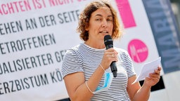 Eine Frau, Maria Mesrian, steht vor einem Plakat und spricht in ein Mikrofon