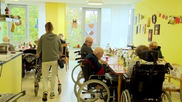 Viele ältere Menschen sitzen in Rollstühlen an mehreren Tischen, eine Person wird von einer Pflegerin geschoben.