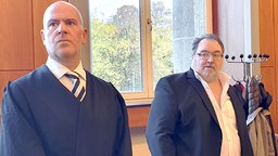 Rechts im Bild steht Michail Ponomarev, ein grauhaariger Mann mit Brille im weißen Hemd und blauen Jackett, links neben ihm sein Verteidiger im blauen Anzug.