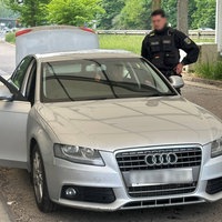 Auf dem Bild ist ein grauer Wagen und mehrere Polizisten. Im Hintergrund ist ein Polizeibulli.