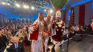 Das Kölner Dreigestirn steht auf einer Bühne und schaut in Richtung der Kamera. Im Hintergrund sitzt das Publikum. 