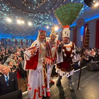 Das Kölner Dreigestirn steht auf einer Bühne und schaut in Richtung der Kamera. Im Hintergrund sitzt das Publikum. 