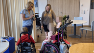 Zwei Frauen stehen in einem Raum. Vor ihnen sind drei Kinder in Rollstühlen zu sehen.
