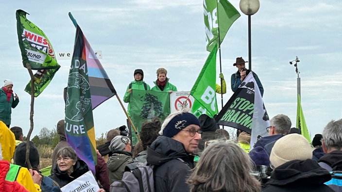 Draußen vor einer kleinen Tribüne stehen Menschen, mit Fahnen. Auf der Tribüne stehen zwei Personen die ein Banner halten. Eine dritte Person spricht in ein Megafone.