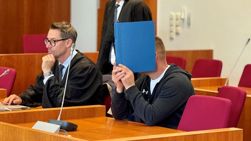 Der Angeklagte und sein Verteidiger sitzen in einem Gerichtssaal. Der Angeklagte verdeckt mit einer blauen Mappe sein Gesicht.