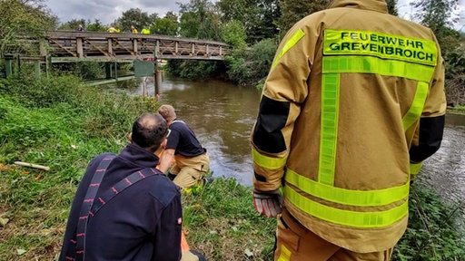 Mehrere Männer an einem Fluss, einer von ihnen trägt eine Jacke mit der Aufschrift "Feuerwehr Grevenbroich"