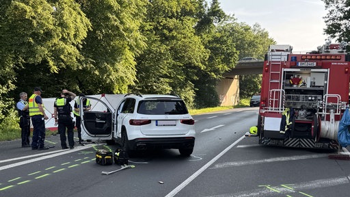 Polizisten stehen an der Unfallstelle neben dem verunfallten weißen Mercedes und einem Feuerwehrauto, auf dem Boden sind gelbe Markierungen des Unfallweges zu sehen