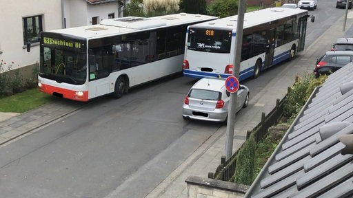Zwei Busse auf einer etwas engeren Straße. Der linke Bus weicht über einen Vorgarten aus. Am Fahrbahnrand parkt ein silberner PKW. Dadurch ist es viel zu eng.