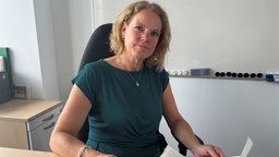 Eine Frau, Ursula Thiel, sitzt in einem Büro und schaut in die Kamera.