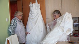 Ein Hochzeitskleid hängt an der Tür, ein anderes wird gerade von einer Dame rechts im Bild begutachtet. Neben ihr ist noch eine andere Frau, die das Kleid an der Tür hält. 