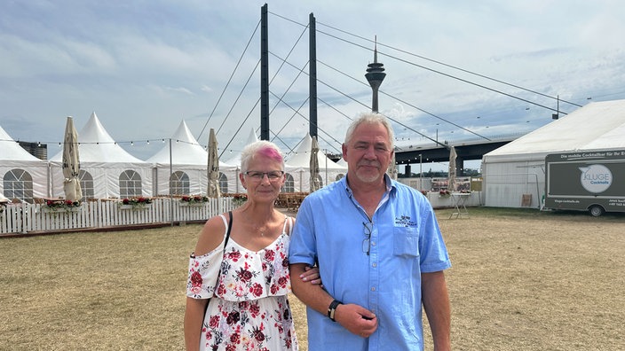 Auf dem Foto sind Astrid Beicht-Schwaab und ihr Ehemann Thomas Schwaab. Die Frau hakt sich bei dem Mann unter. Im Hintergrund ist der Rheinturm.