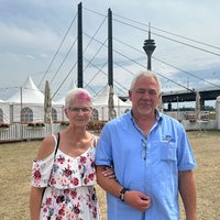 Auf dem Foto sind Astrid Beicht-Schwaab und ihr Ehemann Thomas Schwaab. Die Frau hakt sich bei dem Mann unter. Im Hintergrund ist der Rheinturm.