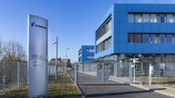 Zukunft zu einem Firmengelände, links eine Säule mit Firmenlogo, hinter einem Tor mehrere blaue Gebäude